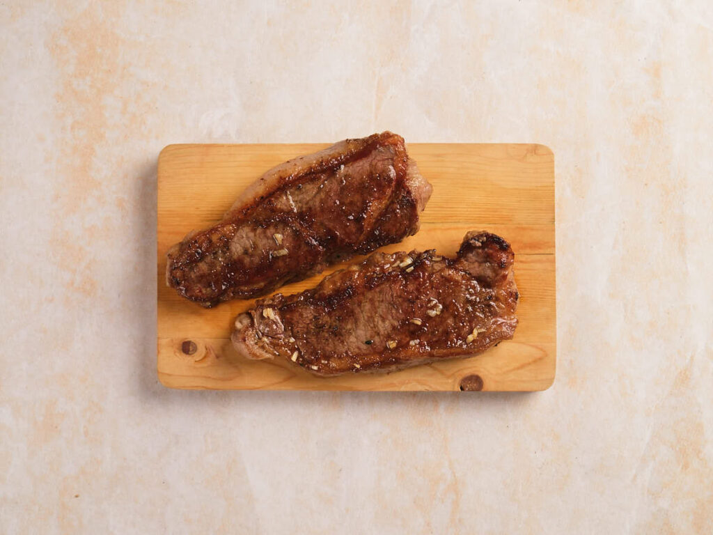Two cooked steak strips with seasoning are placed side by side on a wooden cutting board against a light-colored background.