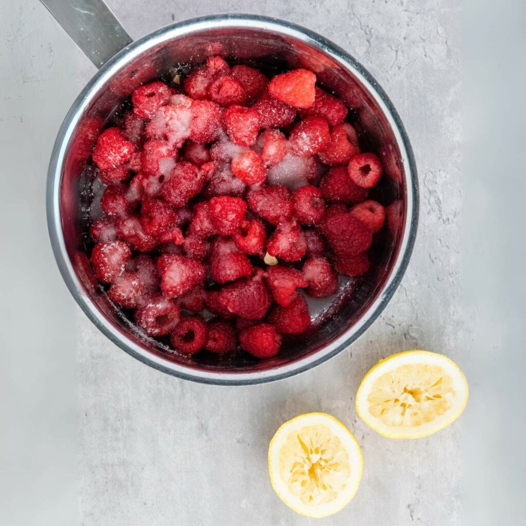 A metal pot filled with raspberries and sugar sits on a gray surface next to two squeezed lemon halves.