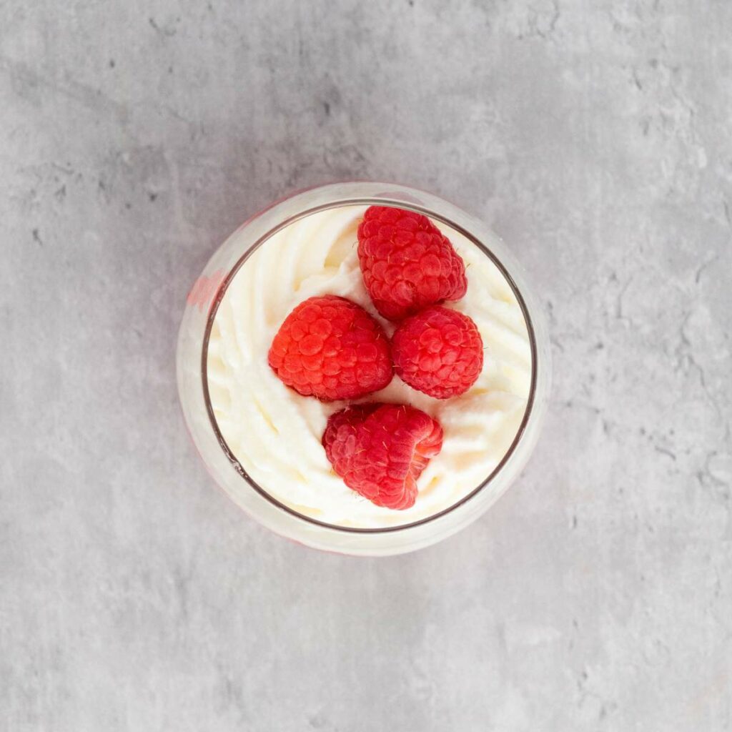 A glass cup filled with whipped cream, topped with four fresh raspberries, on a gray textured surface.