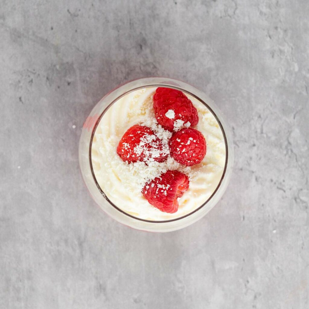 A glass cup filled with whipped cream dessert, topped with three raspberries and a sprinkle of powdered sugar.