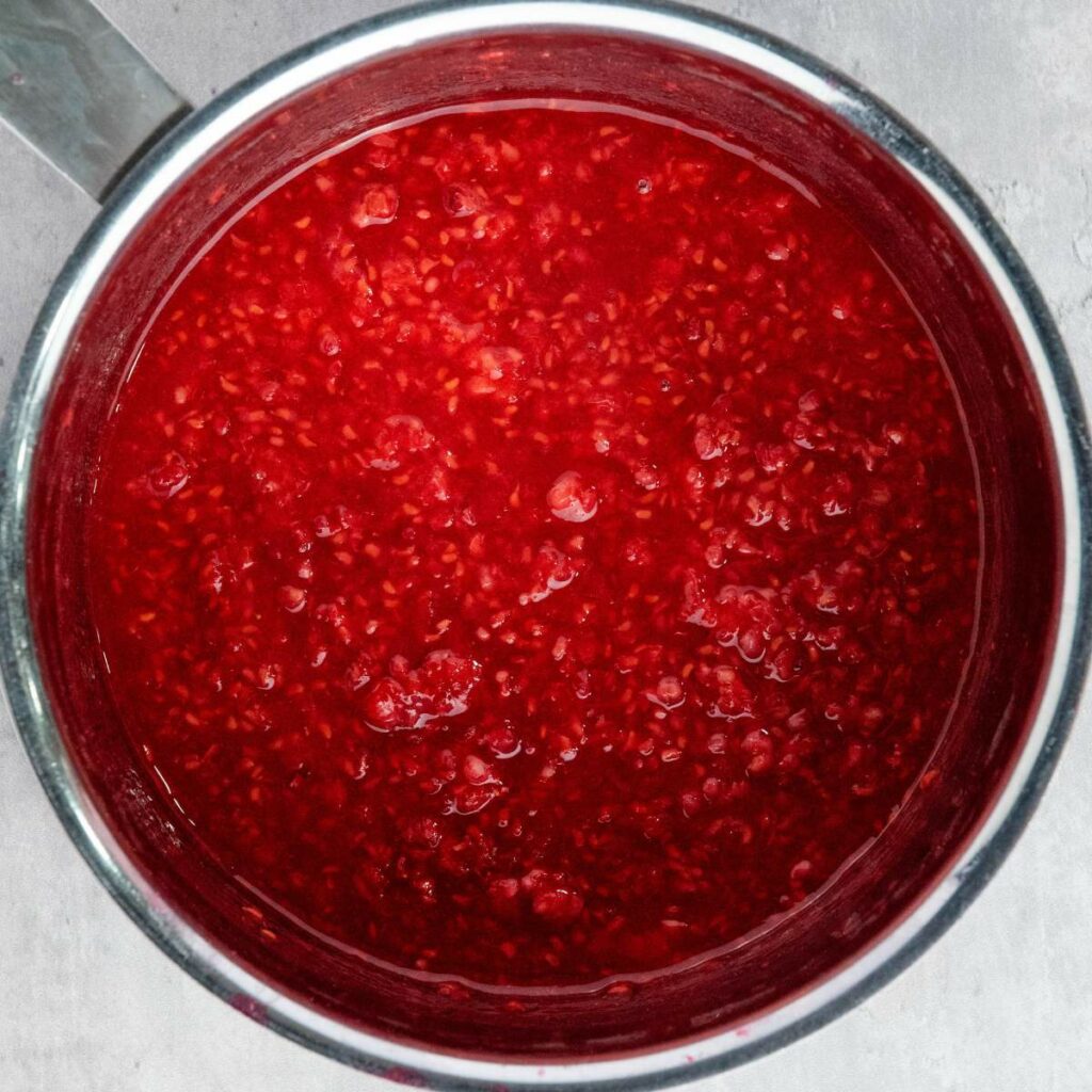 A metal pot filled with bright red raspberry sauce with visible seeds, shown from above on a light gray surface.