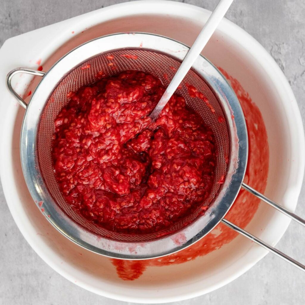 A metal strainer with mashed raspberries sits over a white bowl, with a spoon resting inside the strainer.