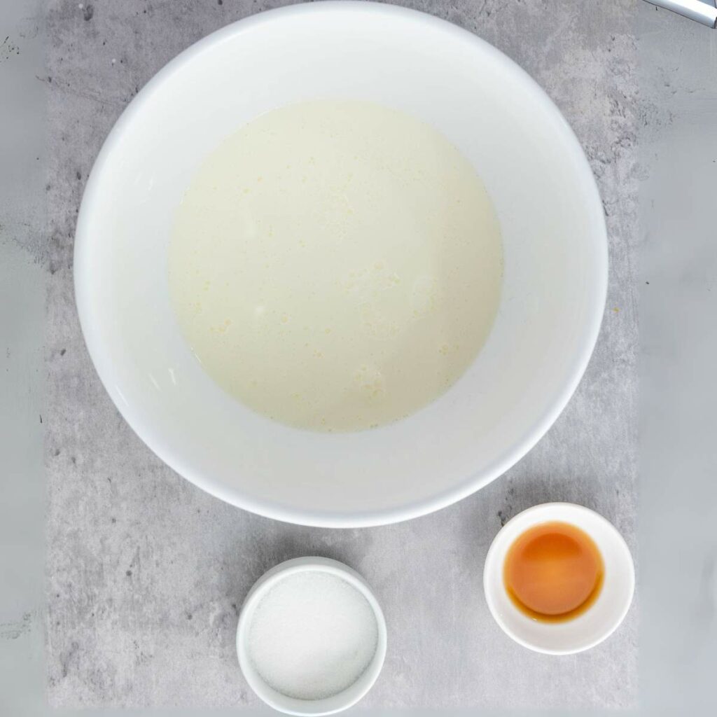 A white bowl with milk, a small bowl of sugar, and a small bowl of vanilla extract on a gray surface.
