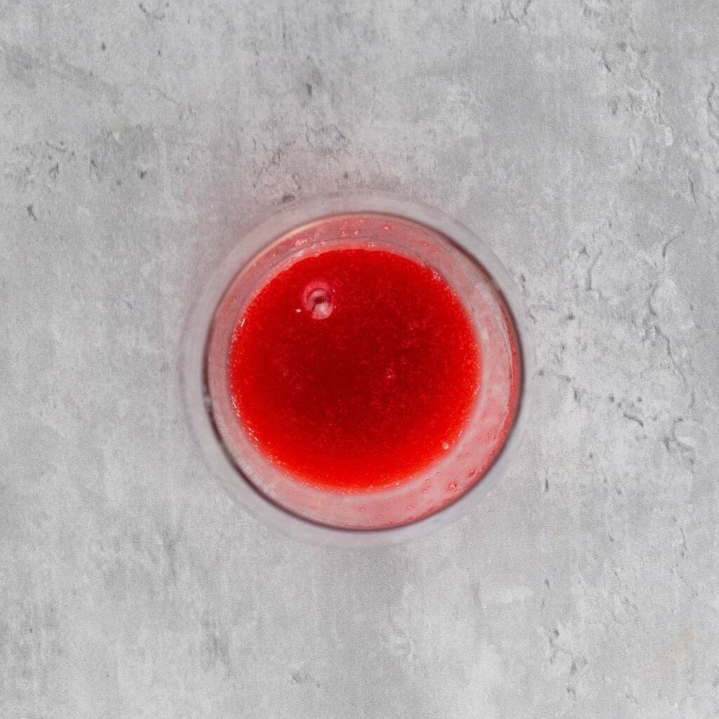 A glass filled with bright red liquid is placed on a light gray textured surface, viewed from above.