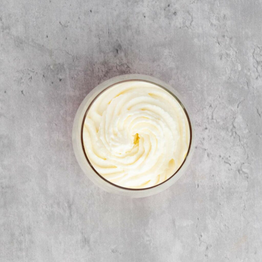 A glass bowl filled with swirled whipped cream sits on a light gray textured surface.