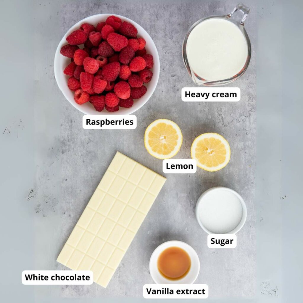 A flat lay of ingredients including raspberries, heavy cream, lemon halves, white chocolate, sugar, and vanilla extract.