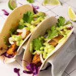 Two soft fish tacos filled with sliced onions, purple cabbage, cilantro, and green sauce.