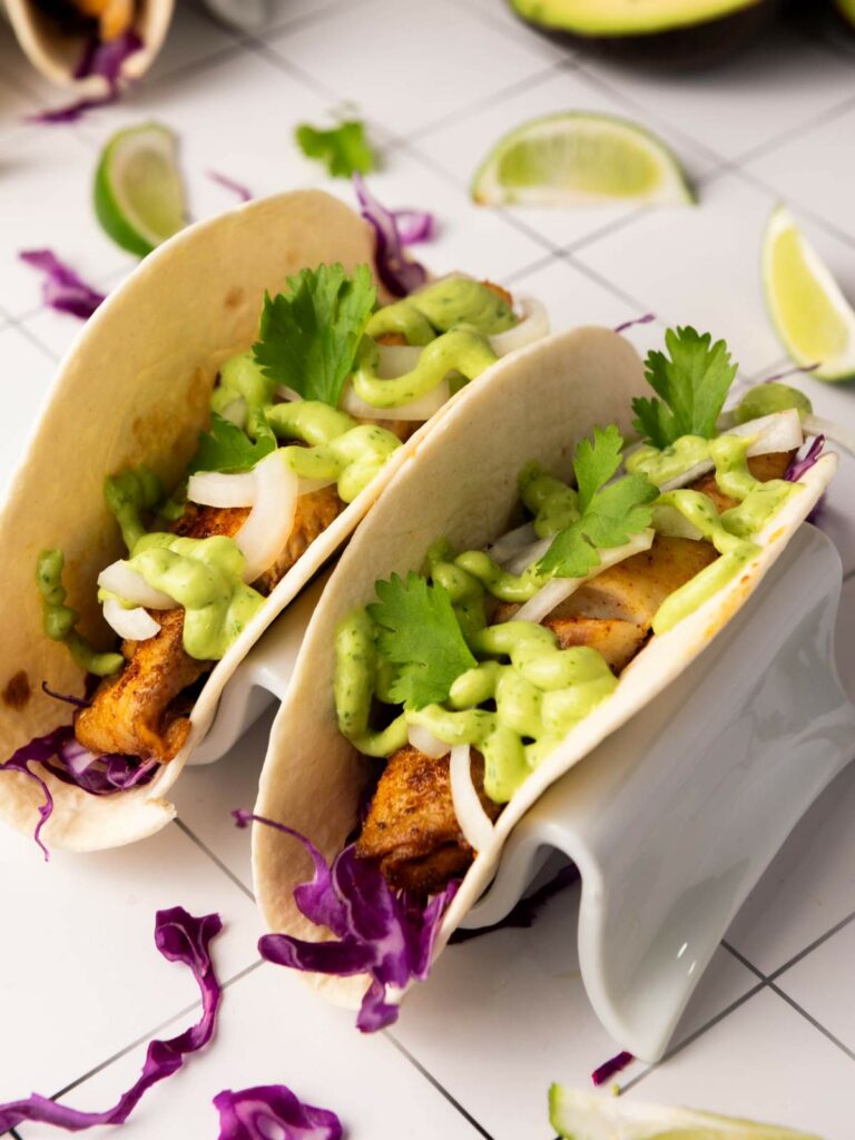 Two soft fish tacos filled with sliced onions, purple cabbage, cilantro, and green sauce.
