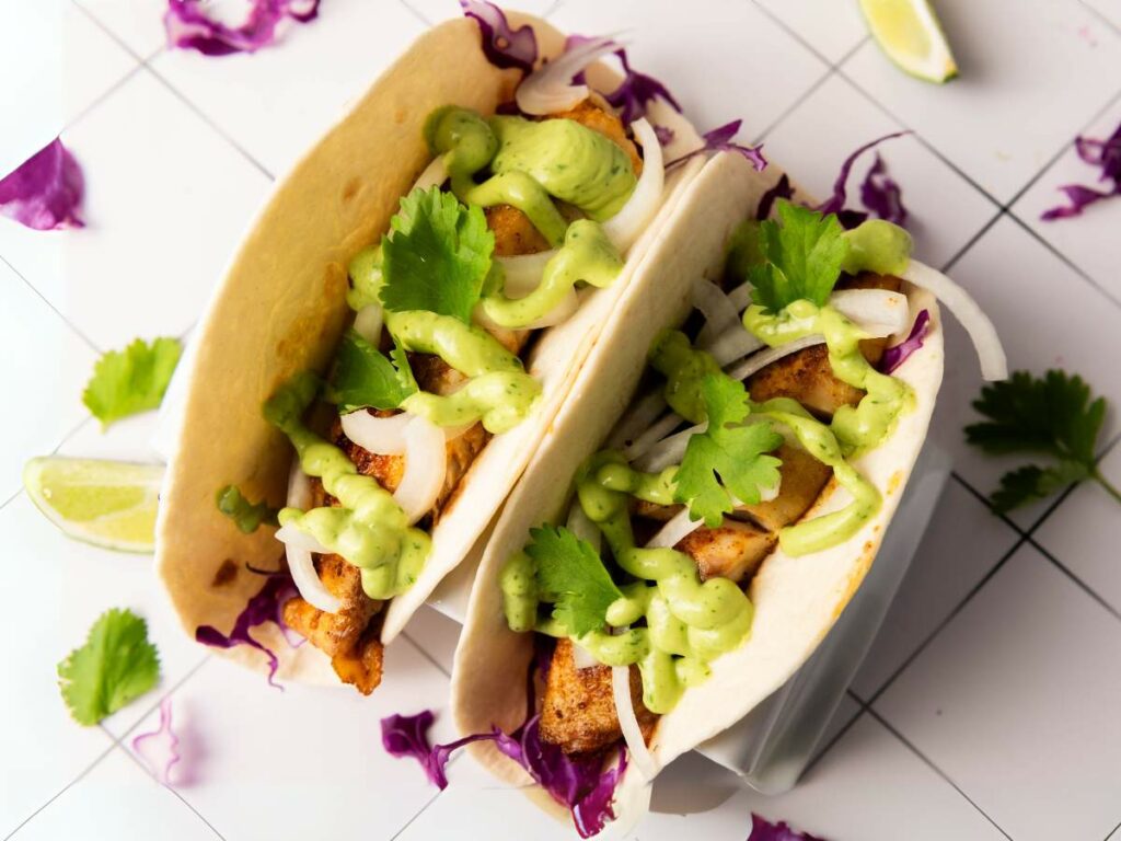 Two soft fish tacos filled with shredded cabbage, onions, cilantro, and drizzled with green sauce.