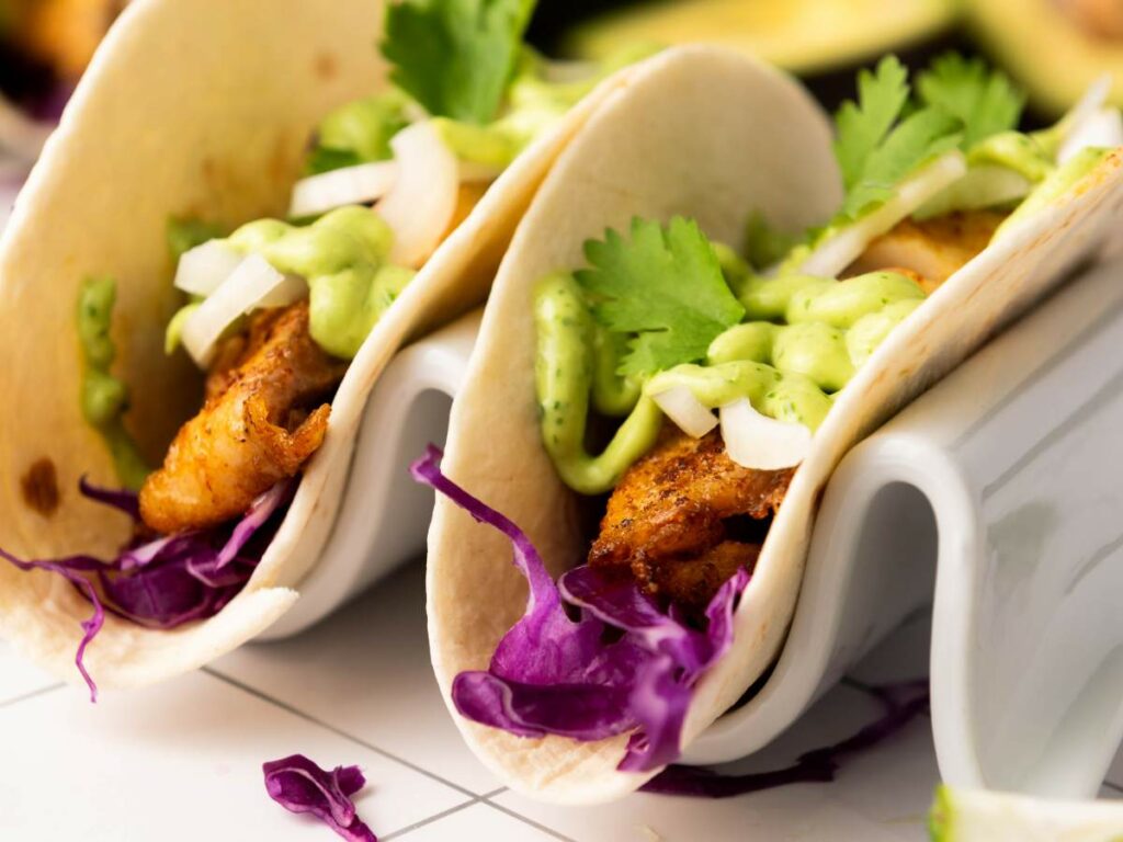 Two soft tacos filled with seasoned grilled fish, shredded purple cabbage, chopped onions, cilantro, and a drizzle of green sauce.
