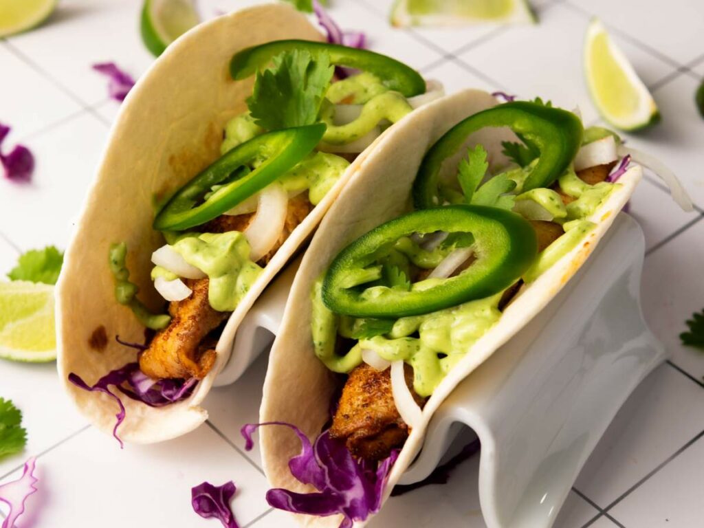 Two fish tacos in a white holder topped with sliced jalapeños, cilantro, onions, green sauce, and purple cabbage.
