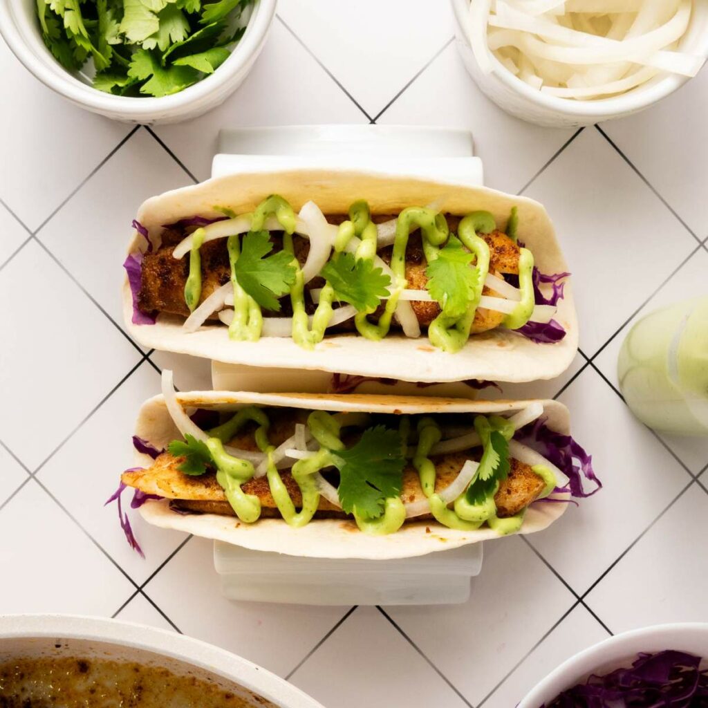 Two fish tacos in soft tortillas topped with sliced onions, green sauce, fresh cilantro, and cabbage.