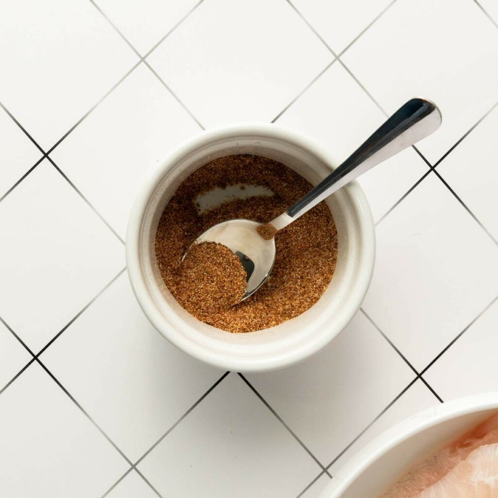 A metal spoon in a white bowl filled with brown spice mix.
