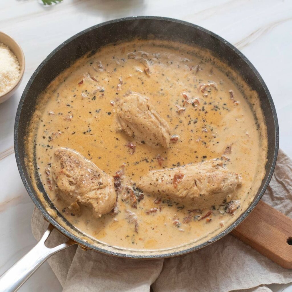 Three pieces of chicken breast simmer in a creamy, herb-seasoned sauce with visible bits of sun-dried tomatoes.