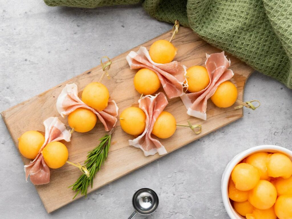 Wooden board with skewers of cantaloupe balls and prosciutto, garnished with a rosemary sprig.