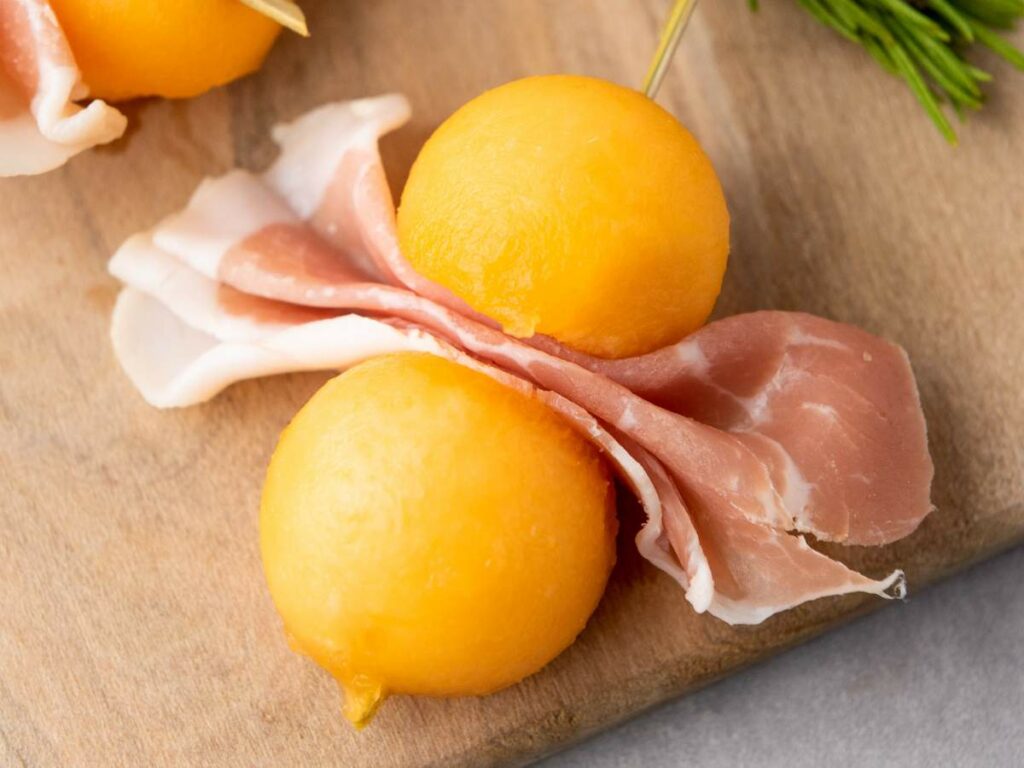 Two cantaloupe melon balls and a slice of prosciutto are skewered together on a wooden stick.