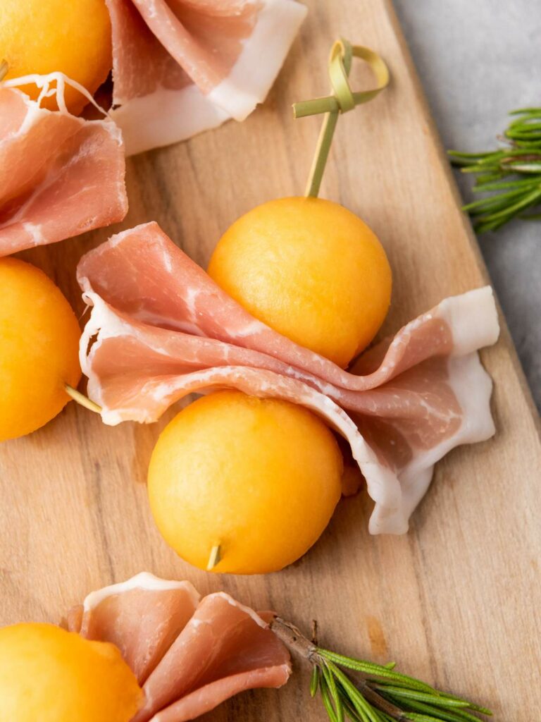 Slices of prosciutto and yellow melon balls are skewered on wooden sticks.