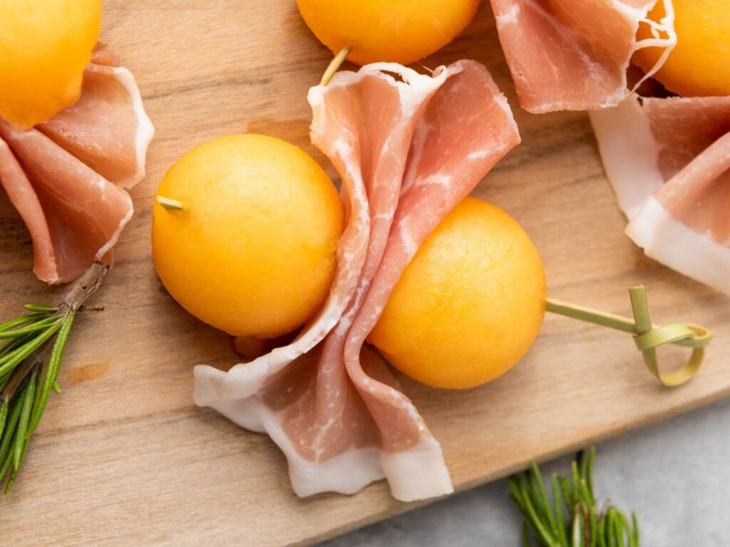 Two yellow melon balls wrapped with thin slices of prosciutto, held together by a skewer.