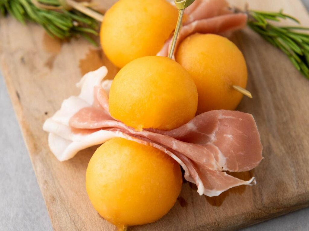 Skewer with slices of prosciutto and yellow melon balls on a wooden board, with a sprig of rosemary in the background.