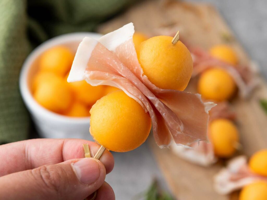 A hand holds a skewer with two cantaloupe balls and a slice of prosciutto; more skewers and a bowl of cantaloupe are in the background.