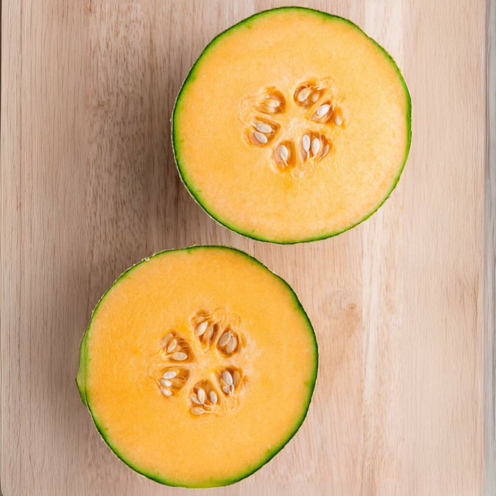 A cantaloupe cut in half, showing orange flesh and seeds, placed on a wooden surface.