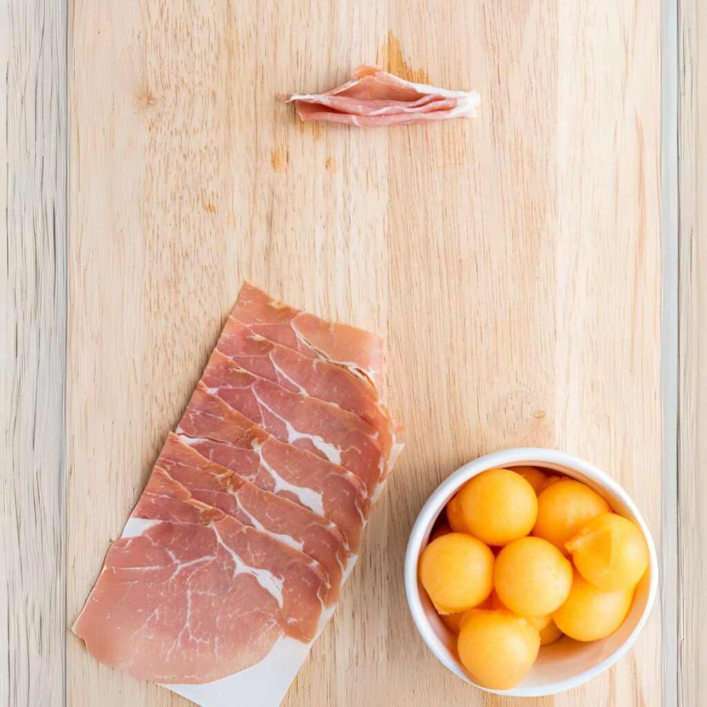 Several slices of prosciutto, a small bowl of cantaloupe balls, and one rolled prosciutto slice.