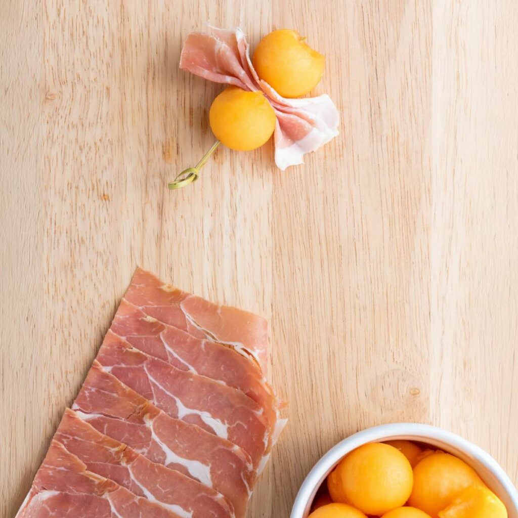 Slices of prosciutto, melon balls in a bowl, and a skewer with prosciutto and melon balls arranged on a wooden surface.