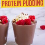 Two glasses of chocolate protein pudding topped with whipped cream, raspberries, and chocolate shavings on a marble surface.