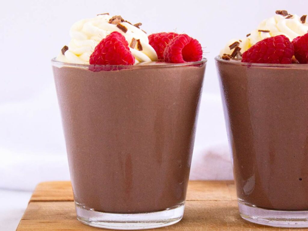 A glass of chocolate mousse topped with whipped cream, fresh raspberries, and chocolate shavings sits on a wooden surface.