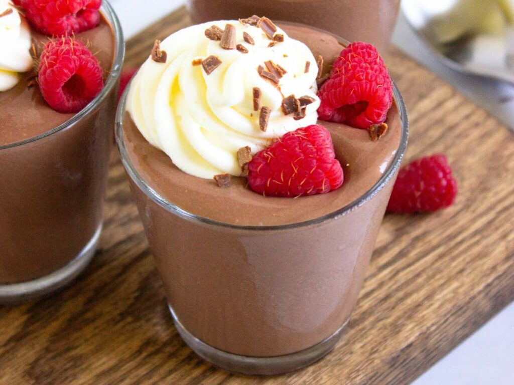 A glass of chocolate mousse topped with whipped cream, chocolate shavings, and fresh raspberries on a wooden surface.