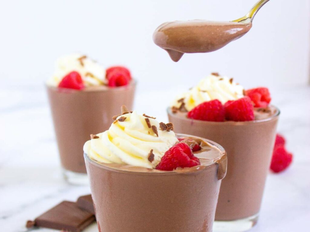 Three glasses of chocolate mousse topped with whipped cream, chocolate shavings, and raspberries; a spoonful of mousse is held above one glass.