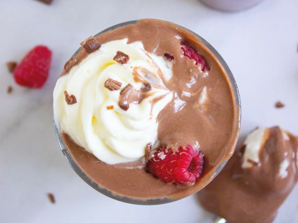 A glass of chocolate mousse topped with whipped cream, chocolate shavings, and raspberries on a white surface.