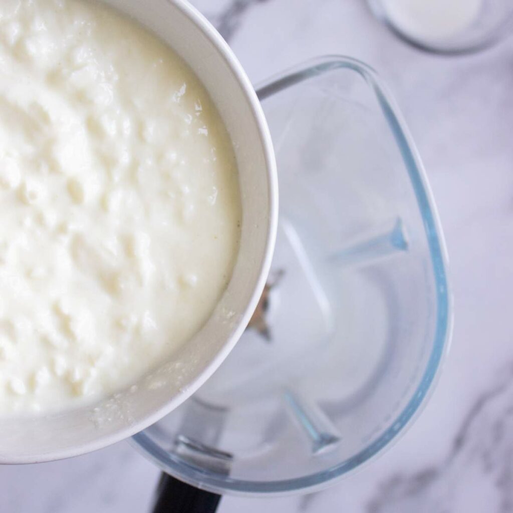 A bowl of curdled milk is being poured into a blender on a marble countertop.