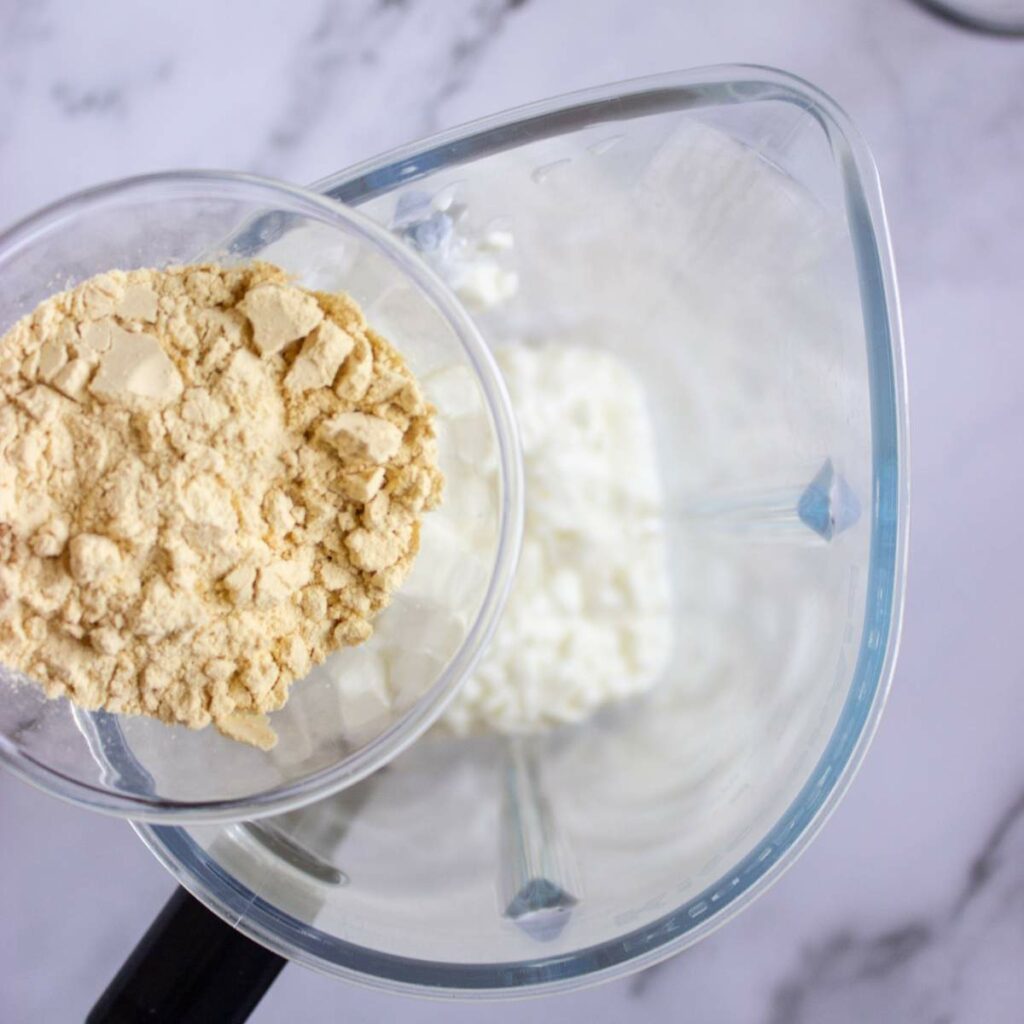 A measuring cup with protein powder is being poured into a blender containing cottage cheese on a marble countertop.