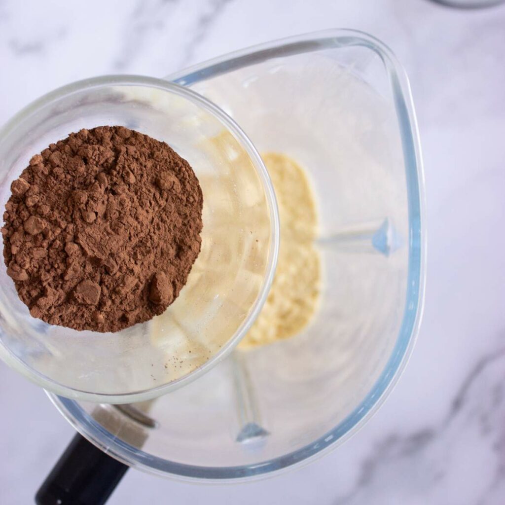 A glass filled with cocoa powder is being held above a blender containing a yellow liquid mixture.