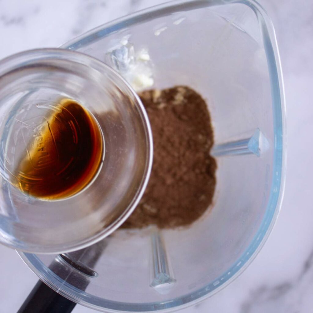 A glass container with vanilla extract is being poured into a blender containing cocoa powder.