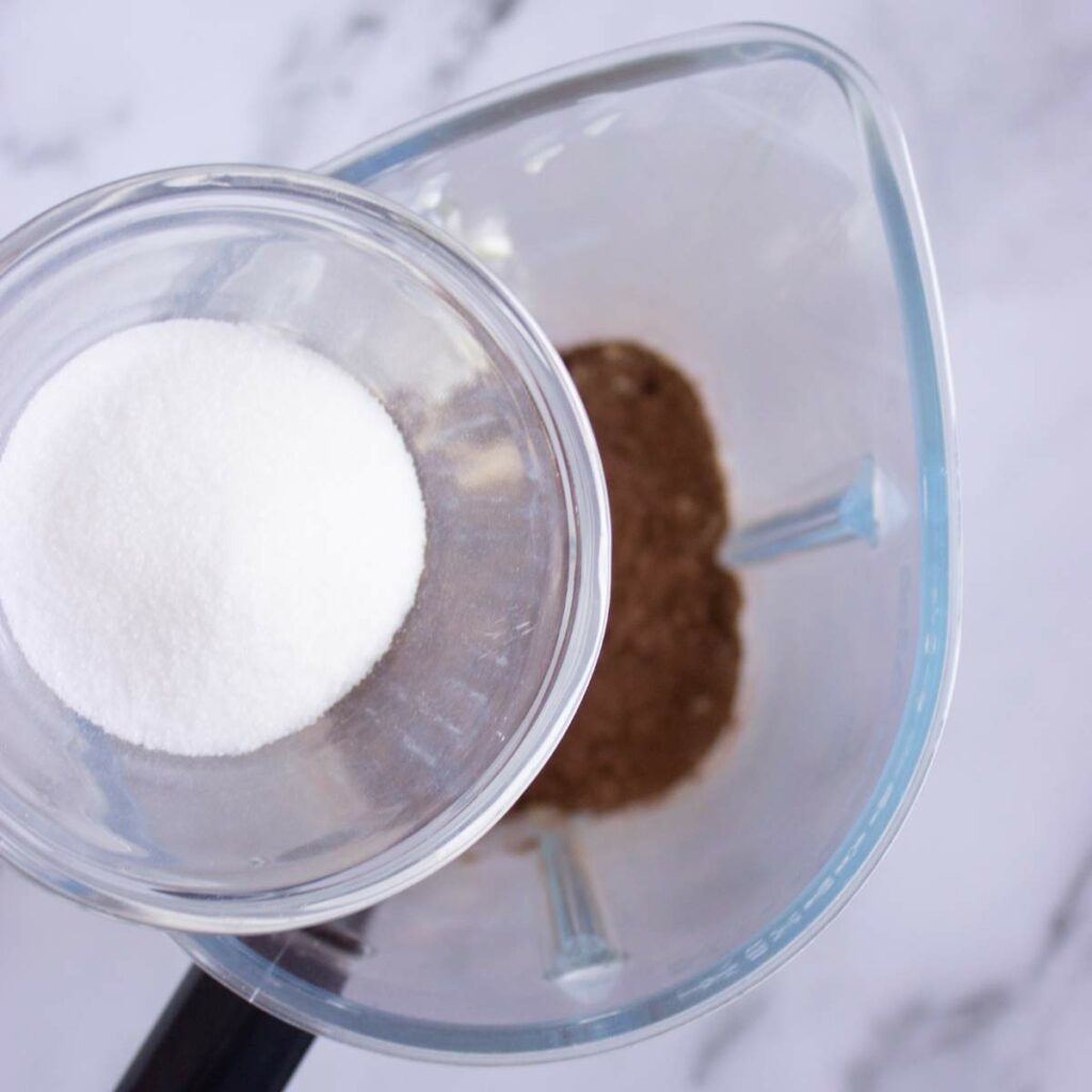 A glass bowl of granulated sugar is being poured into a blender containing cocoa powder.