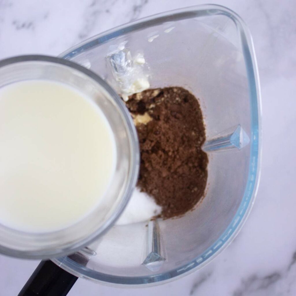 A blender containing milk, cocoa powder, and a white substance, possibly sugar or protein powder, viewed from above on a marble surface.