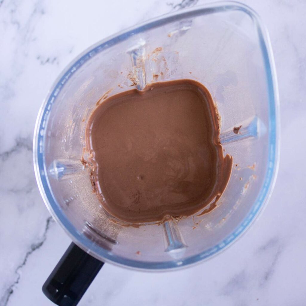 A blender containing a small amount of chocolate liquid mixture sits on a white marble surface.