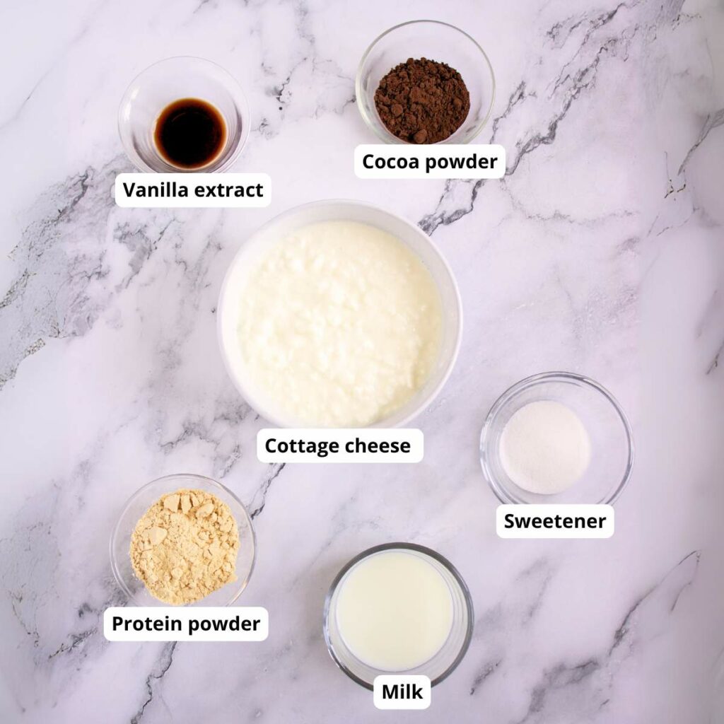 Six labeled bowls on a marble surface containing vanilla extract, cocoa powder, cottage cheese, sweetener, protein powder, and milk.