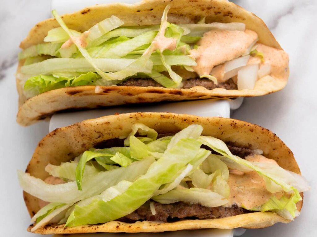 Two soft-shell smash burger taco filled with ground beef, shredded lettuce, chopped onions, and a creamy orange sauce.