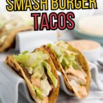 Two smash burger tacos in taco holders are filled with ground beef, shredded lettuce, and sauce, with more tacos and a dipping sauce in the blurred background.