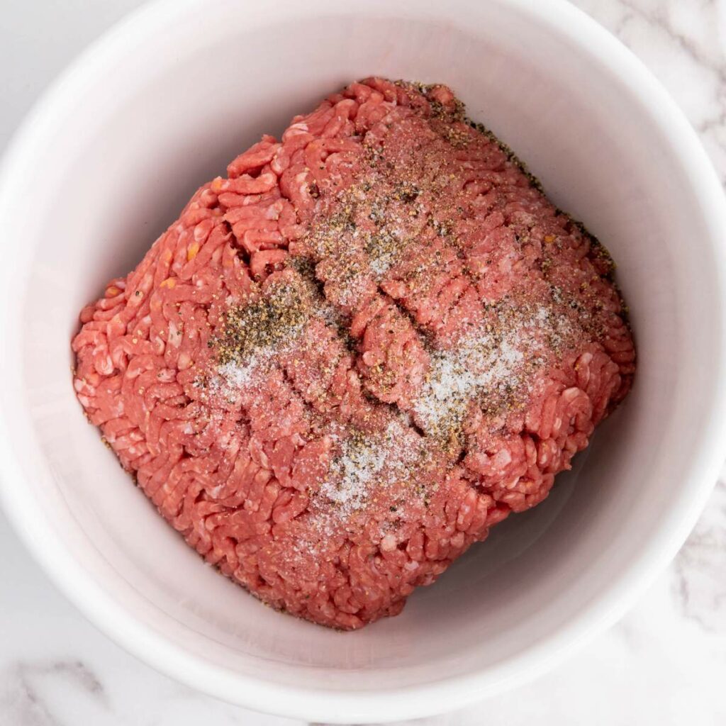 Raw ground beef in a white bowl, topped with salt and black pepper.
