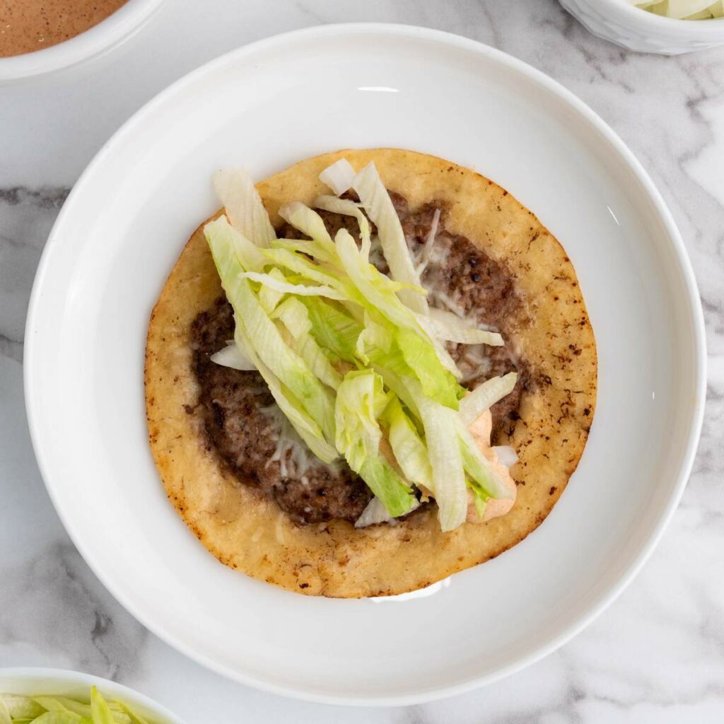 A flat, fried tortilla topped with seasoned ground beef, shredded lettuce, and melted cheese.