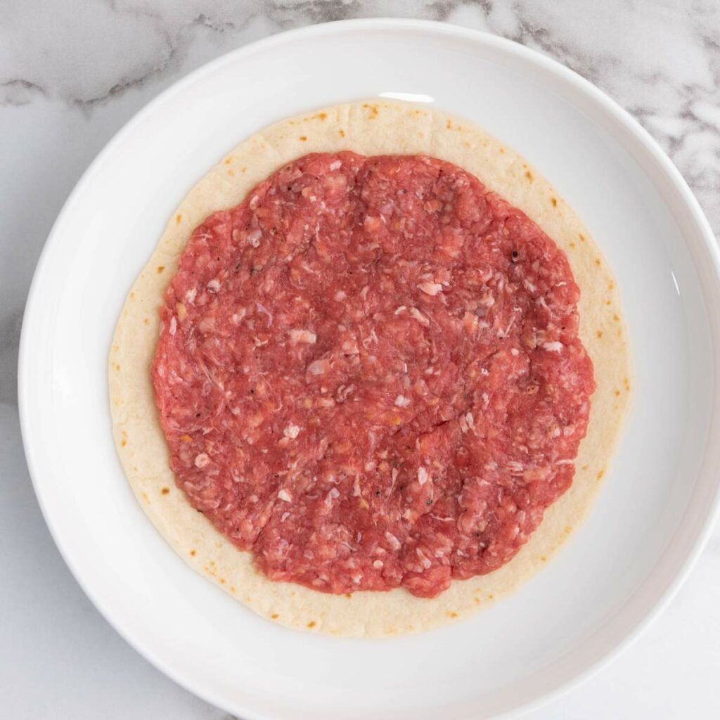 Raw ground meat is spread in a circular shape on top of a tortilla.