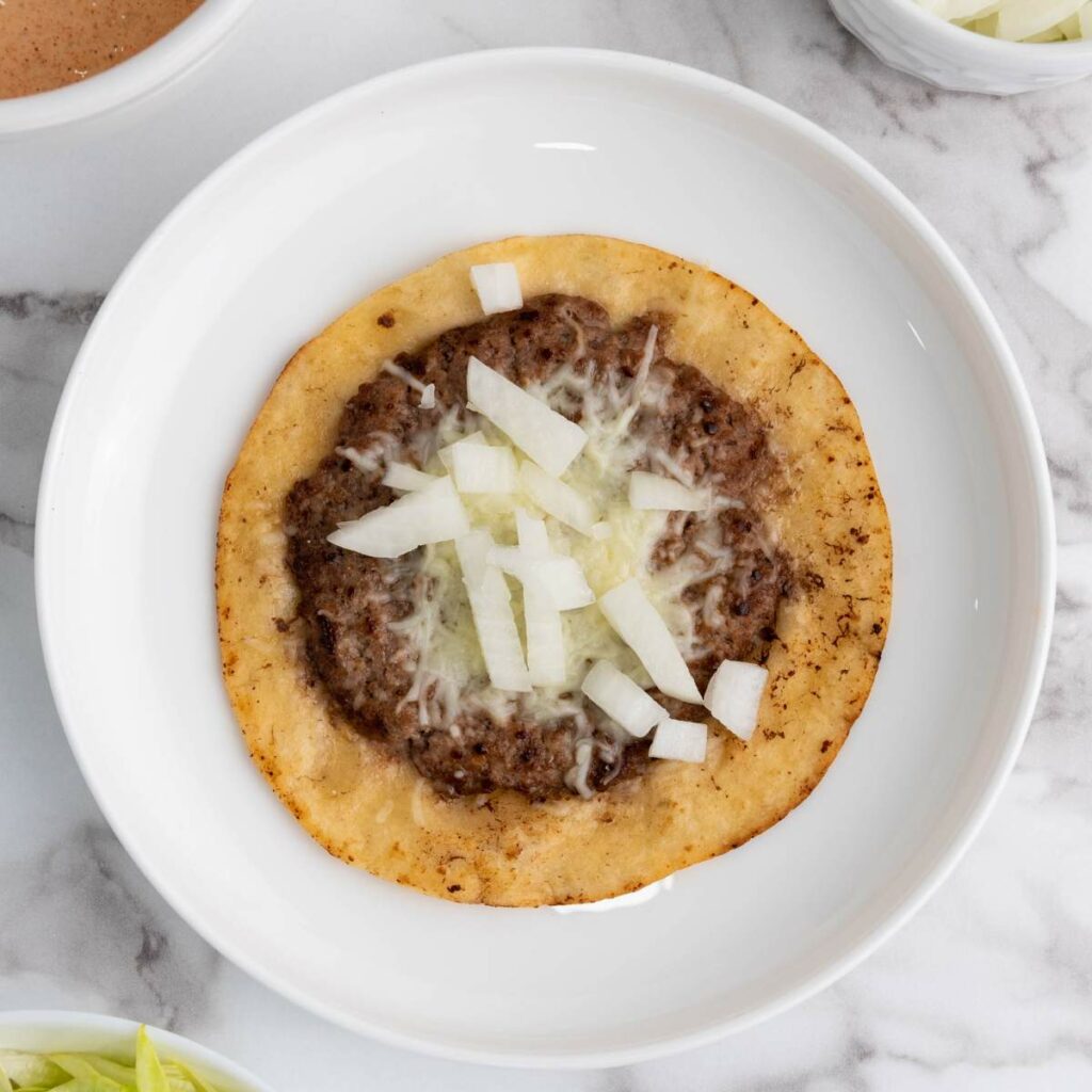 A flat, round taco with ground beef, melted cheese, and chopped raw onions.
