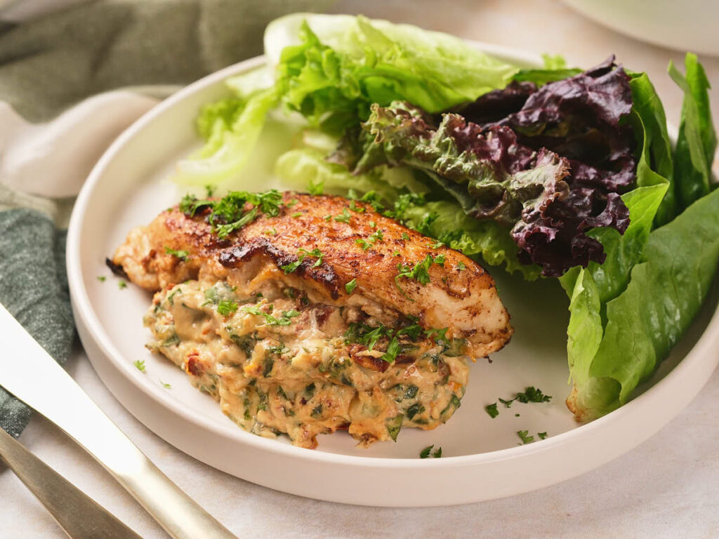Grilled stuffed chicken breast with a creamy herb filling, served with assorted fresh lettuce leaves on a white plate.