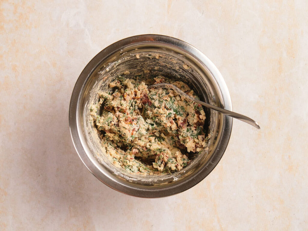 A metal bowl with a spoon holds a mixed blend of chopped herbs, sun-dried tomatoes, and cheese on a light surface.