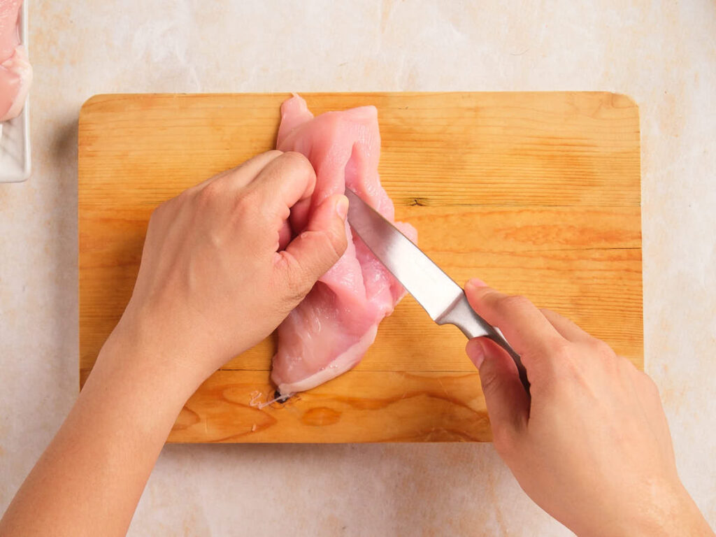 Two hands cutting raw chicken breast on a wooden cutting board with a knife.