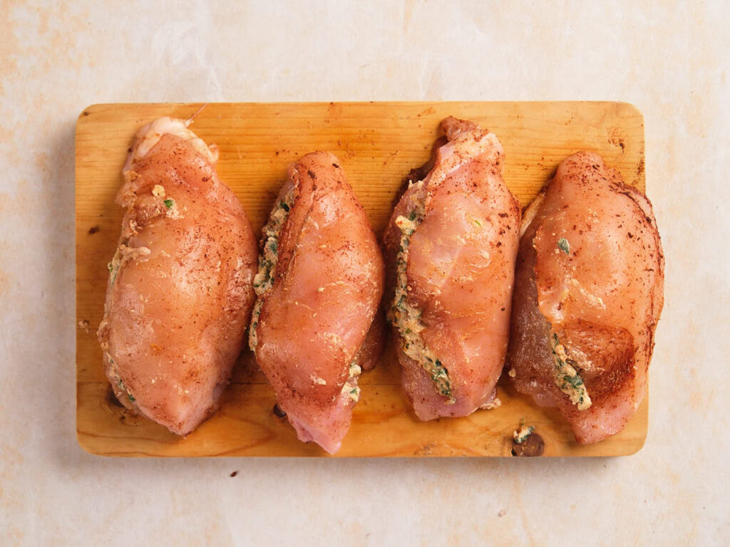 Four raw, seasoned chicken breasts stuffed with a herb and cheese mixture are arranged in a row on a wooden cutting board.